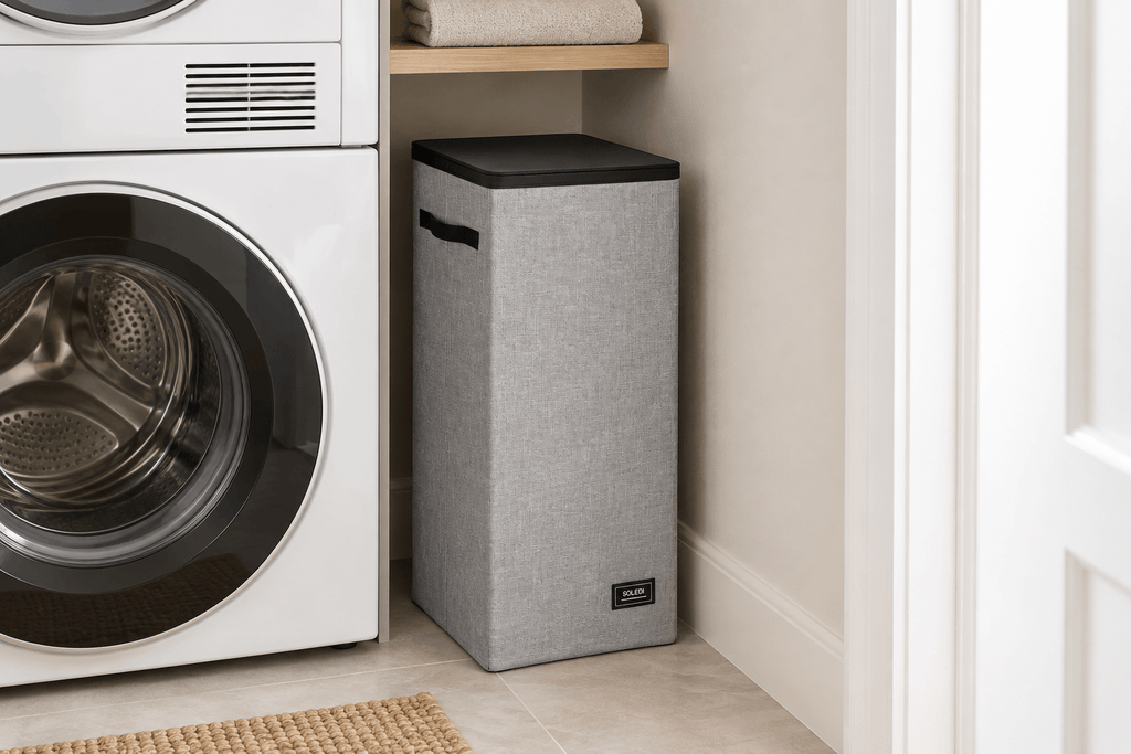 Gray SOLEDI slim laundry hamper with a closed black lid positioned beside a stacked washer and dryer.