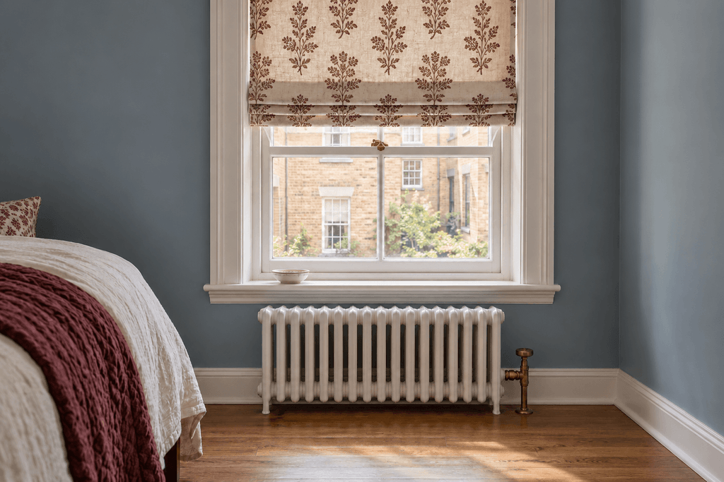 Burgundy botanical Roman shade above a bedroom window, leaving the radiator and its brass valve uncovered.