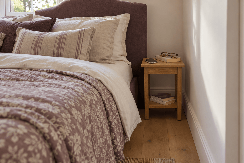 Small wooden bedside table in a narrow gap between the bed and the wall holding a phone, glasses and a paperback beside a bed with layered plum and ivory bedding.
