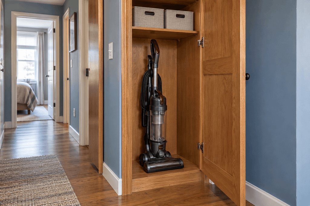 Upright vacuum stored in an open wooden hallway cupboard beneath a shelf with two fabric storage bins.