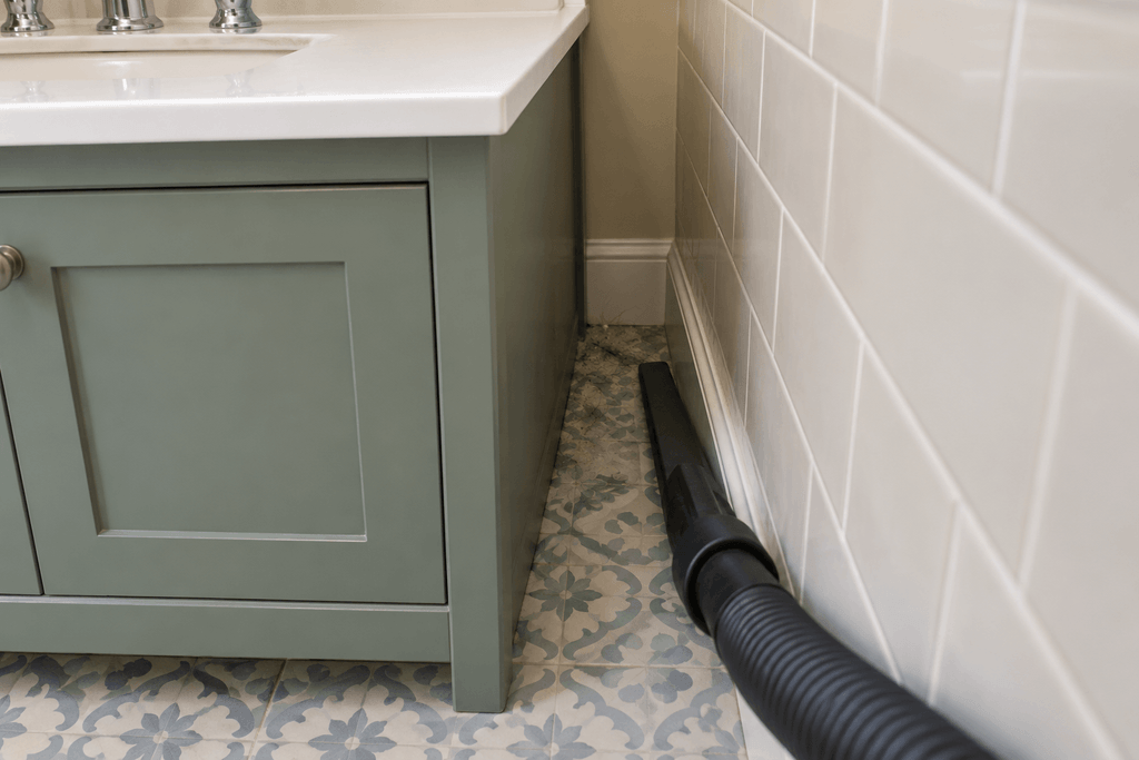 Narrow vacuum attachment reaching along the tiled floor between a green bathroom vanity and the side wall.