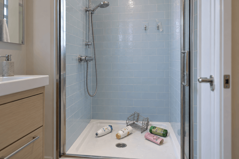 Small shower with peeling adhesive mounts high on the tiled wall and a fallen wire shelf and scattered toiletries on the shower floor.
